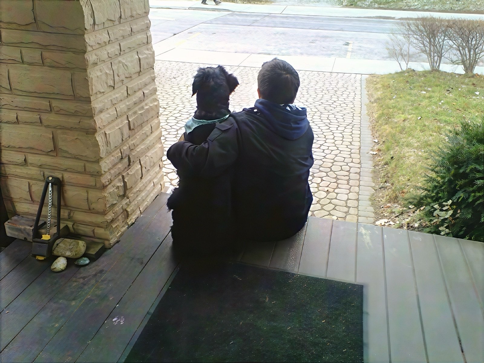 Mark sitting on a front porch with his arm around Riley, a black giant schnauzer wearing a blue bandana